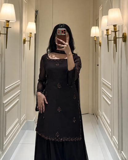 A woman takes a mirror selfie wearing a black embroidered kurta with a matching skirt in a hallway with wall-mounted lamps.