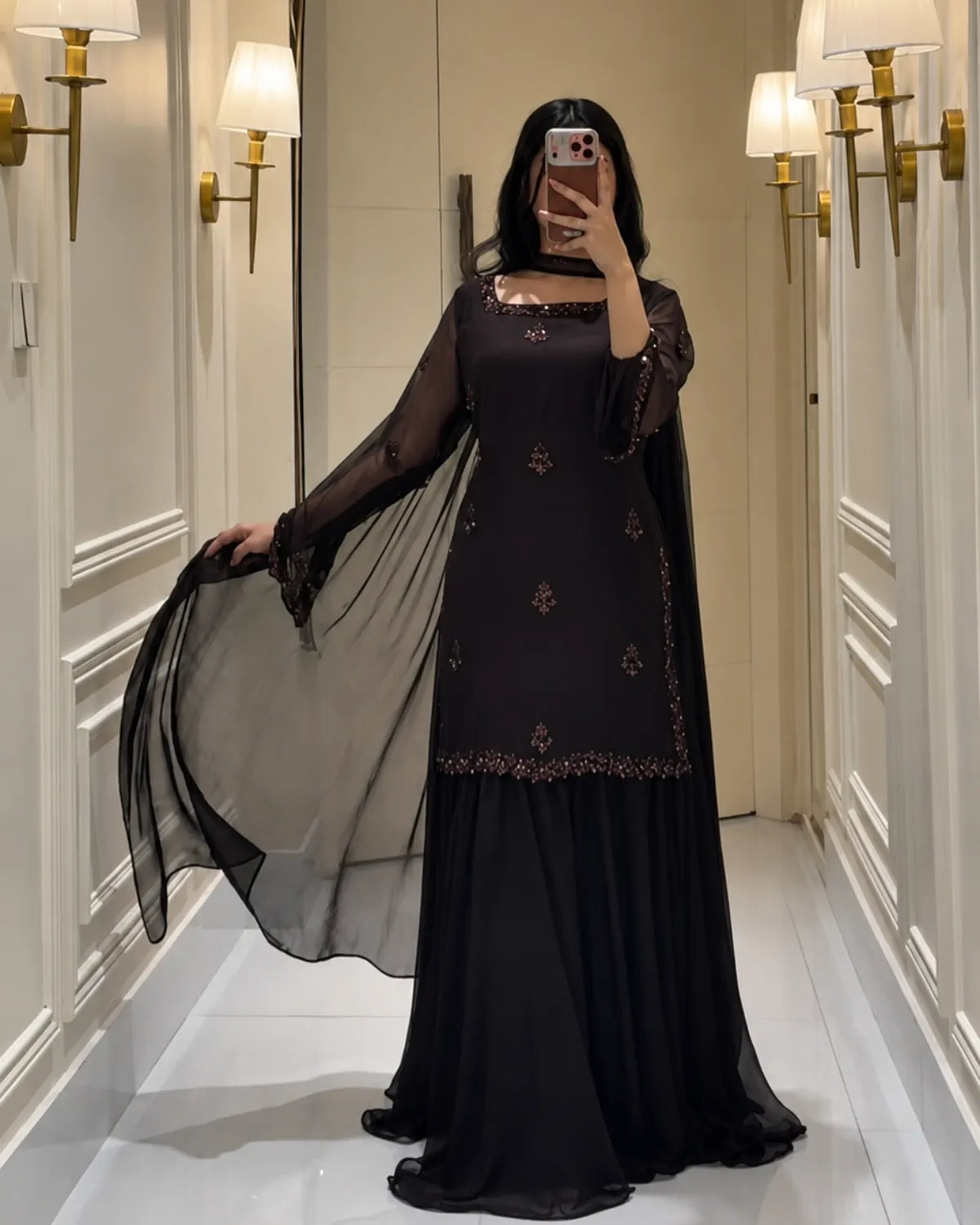 A woman takes a mirror selfie wearing a black long-sleeved dress with gold embroidery and a sheer black dupatta, standing in a hallway with wall sconces.