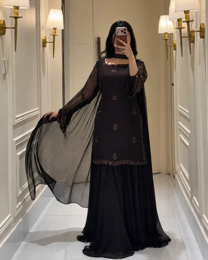 A woman takes a mirror selfie wearing a black long-sleeved dress with gold embroidery and a sheer black dupatta, standing in a hallway with wall sconces.