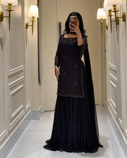 A woman takes a mirror selfie wearing a black traditional outfit with embroidered details and a long sheer dupatta, standing in a hallway with wall sconces.