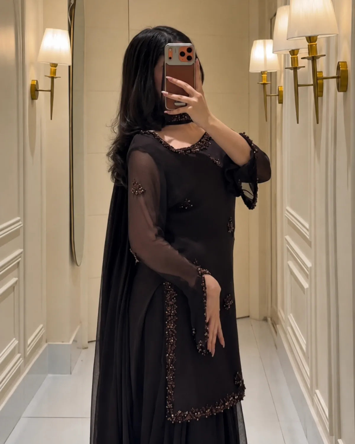 A woman takes a mirror selfie wearing a black long-sleeve kurta with sheer mesh sleeves, a matching draped cape, and intricate dark brown beaded embroidery along the neckline, sleeve cuffs, and hem. She is standing in an elegant hallway with cream paneled walls and gold wall sconces.