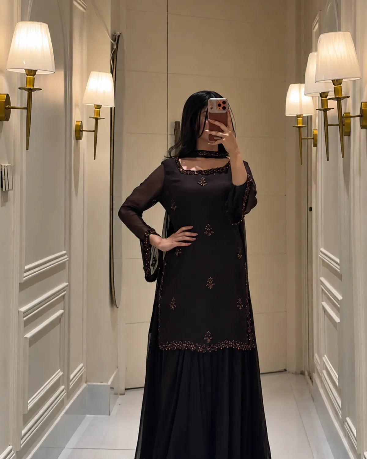 A woman takes a mirror selfie wearing a black long-sleeve kurta with gold embroidery and a matching black skirt, standing in a hallway with wall-mounted lamps.