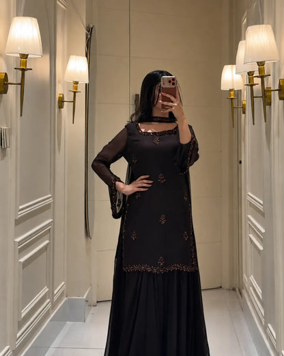 A woman takes a mirror selfie wearing a black long-sleeve kurta with gold embroidery and a matching black skirt, standing in a hallway with wall-mounted lamps.