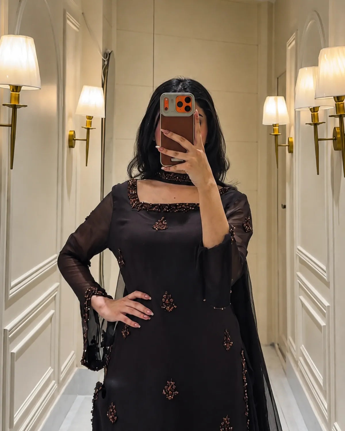 A woman takes a mirror selfie wearing a black traditional outfit with intricate gold and brown embroidery, standing in a hallway with wall sconces.