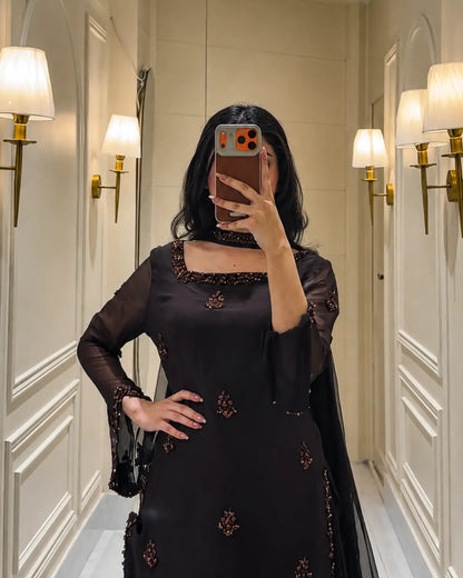 A woman takes a mirror selfie wearing a black traditional outfit with intricate gold and brown embroidery, standing in a hallway with wall sconces.