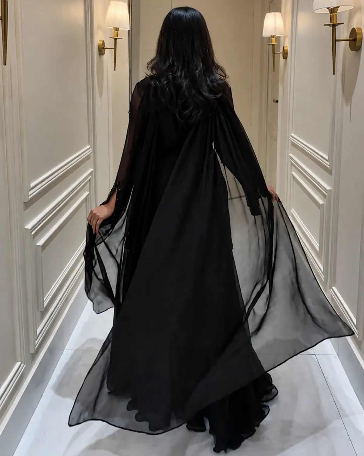 A woman viewed from behind wearing a flowing black sheer maxi dress or robe with long sleeves, standing in a hallway with cream paneled walls and wall sconces.