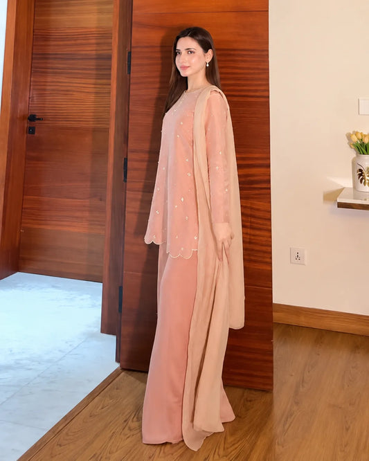 A woman models a dusty pink traditional outfit consisting of a long kurta, matching trousers, and a sheer embroidered dupatta, standing in a room with wooden doors and flooring.