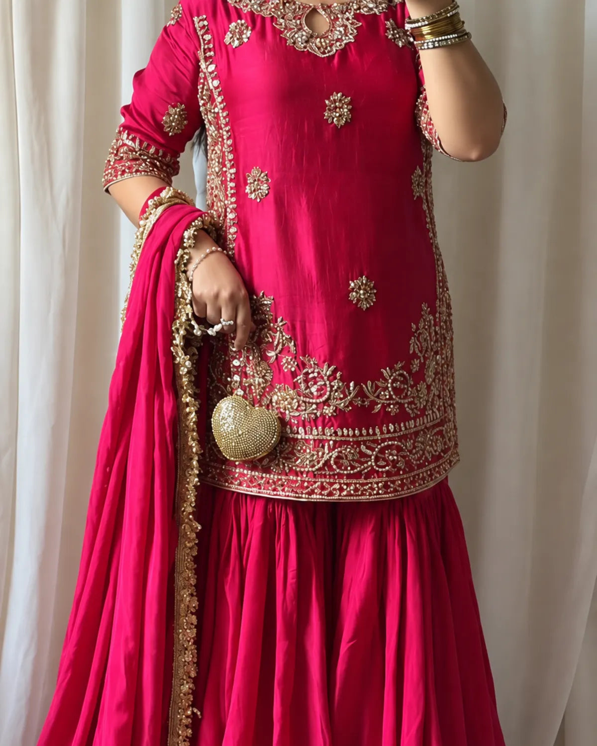A woman wearing a bright pink traditional outfit with ornate gold embroidery, a matching pink dupatta, and gold jewelry, standing against a white curtain backdrop.