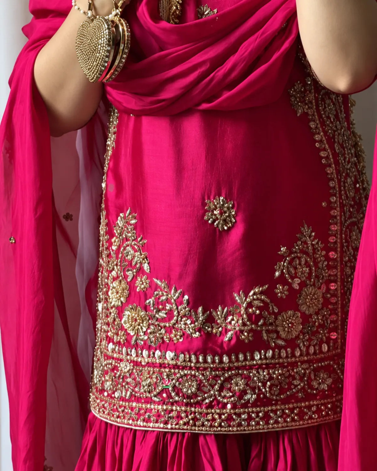 A close-up of a bright pink traditional outfit with extensive gold embroidery and embellishments, featuring a draped dupatta and ornate jewelry.