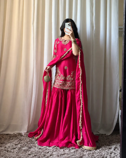 A woman takes a mirror selfie wearing a bright magenta pink traditional outfit with gold embroidery and a matching draped scarf. The outfit is styled with a long skirt and a heavily embellished top, set against a background of cream-colored curtains and a gray shag rug.