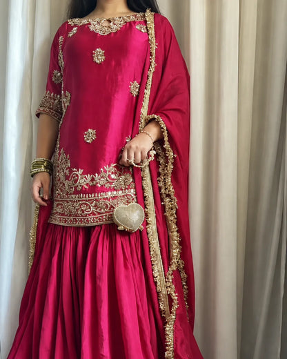 A woman wearing a bright magenta pink traditional outfit with ornate gold embroidery, featuring a kurta, flared skirt, and matching dupatta, standing in front of beige curtains.