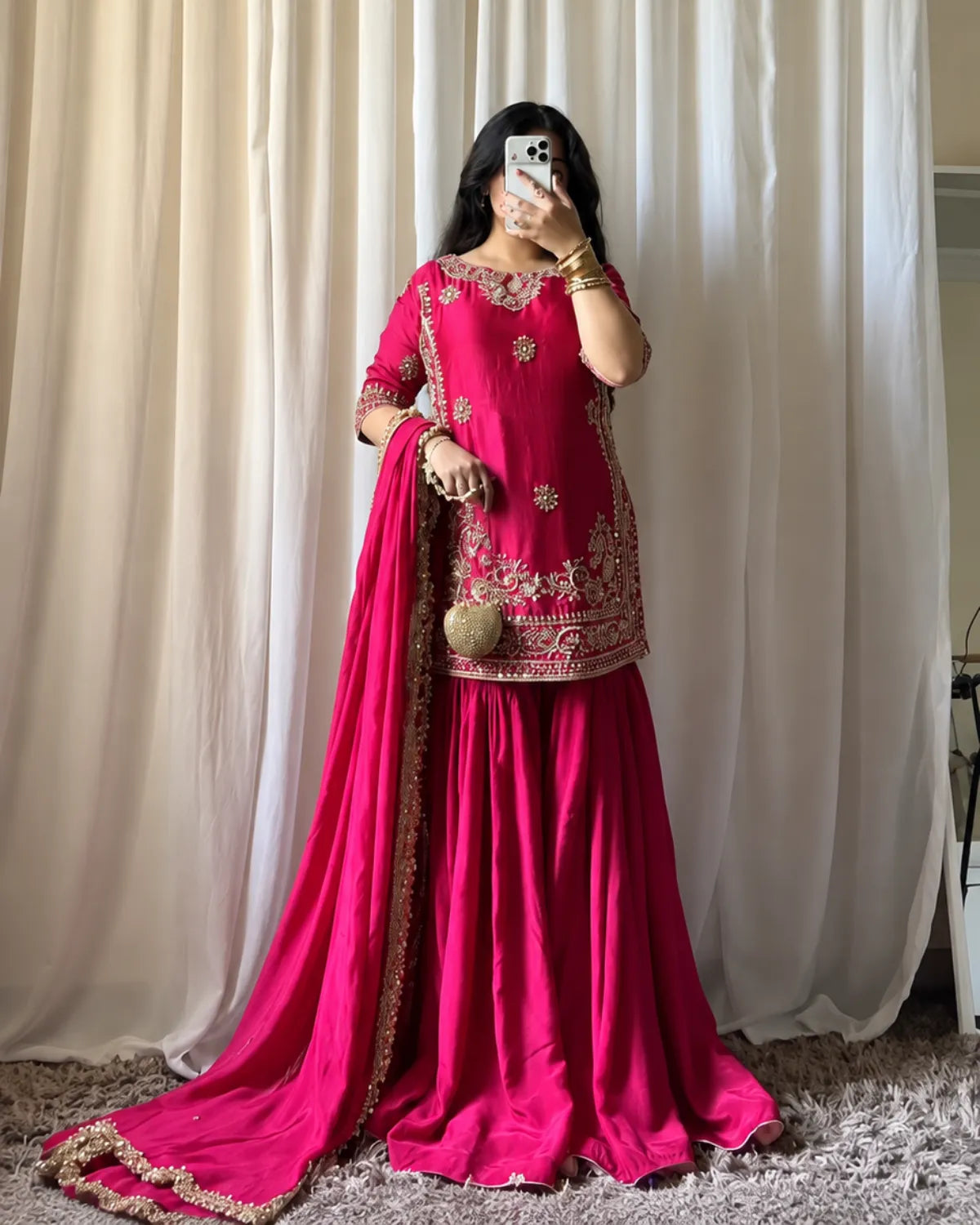 A woman takes a mirror selfie wearing a bright magenta pink traditional Indian outfit with gold embroidery, featuring a kurta, flared skirt, and a matching dupatta draped over the floor.