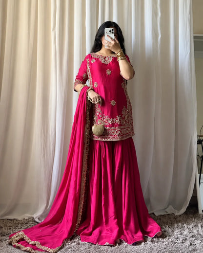 A woman takes a mirror selfie wearing a bright magenta pink traditional Indian outfit with gold embroidery, featuring a kurta, flared skirt, and a matching dupatta draped over the floor.