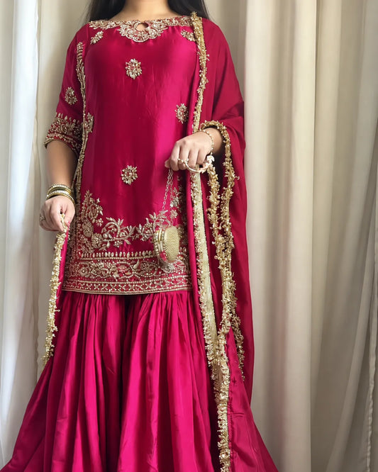 A woman wearing a bright fuchsia pink traditional outfit with ornate gold embroidery and a matching sheer dupatta draped over her shoulders, standing in front of beige curtains.