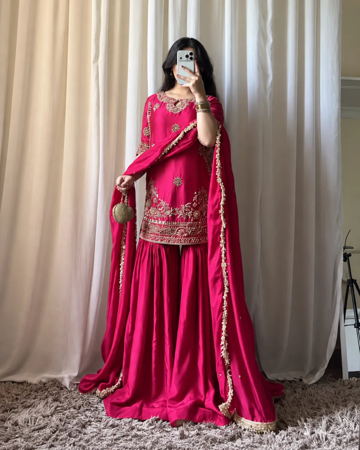 A woman takes a mirror selfie wearing a vibrant magenta pink traditional Indian outfit with gold embroidery, a matching dupatta, and a small gold handbag, set against a backdrop of cream curtains and a shag rug.