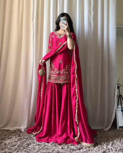 A woman takes a mirror selfie wearing a vibrant magenta pink traditional Indian outfit with gold embroidery, a matching dupatta, and a small gold handbag, set against a backdrop of cream curtains and a shag rug.