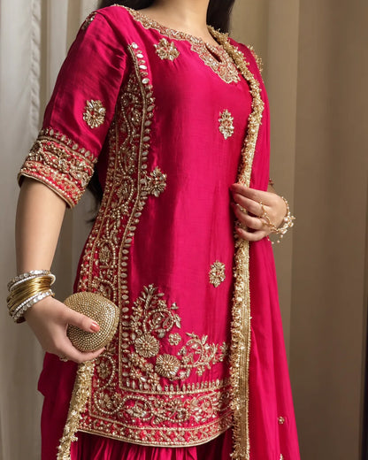 A woman wearing a bright pink traditional outfit with extensive gold embroidery and a matching pink dupatta, accessorized with gold bangles and a gold clutch.