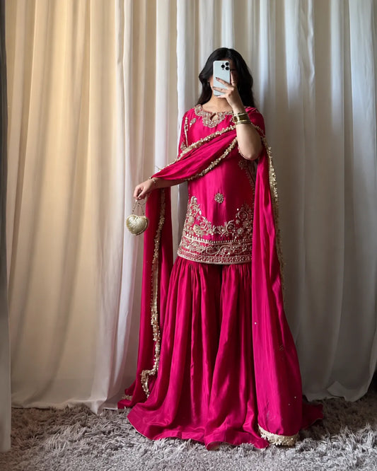 A woman takes a mirror selfie wearing a bright fuchsia pink traditional Indian salwar kameez outfit with heavy gold embroidery and a matching dupatta draped over her shoulder. She is standing in front of beige curtains on a gray shag rug.