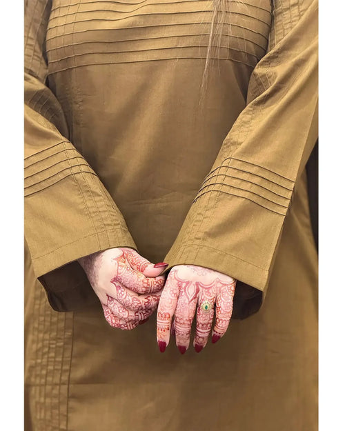 A close-up of a person wearing a tan long-sleeved garment with horizontal pleats on the chest and cuffs, showing both hands decorated with intricate red henna patterns and a small green ring.