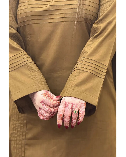 A close-up of a person wearing a tan long-sleeved garment with horizontal pleats on the chest and cuffs, showing both hands decorated with intricate red henna patterns and a small green ring.