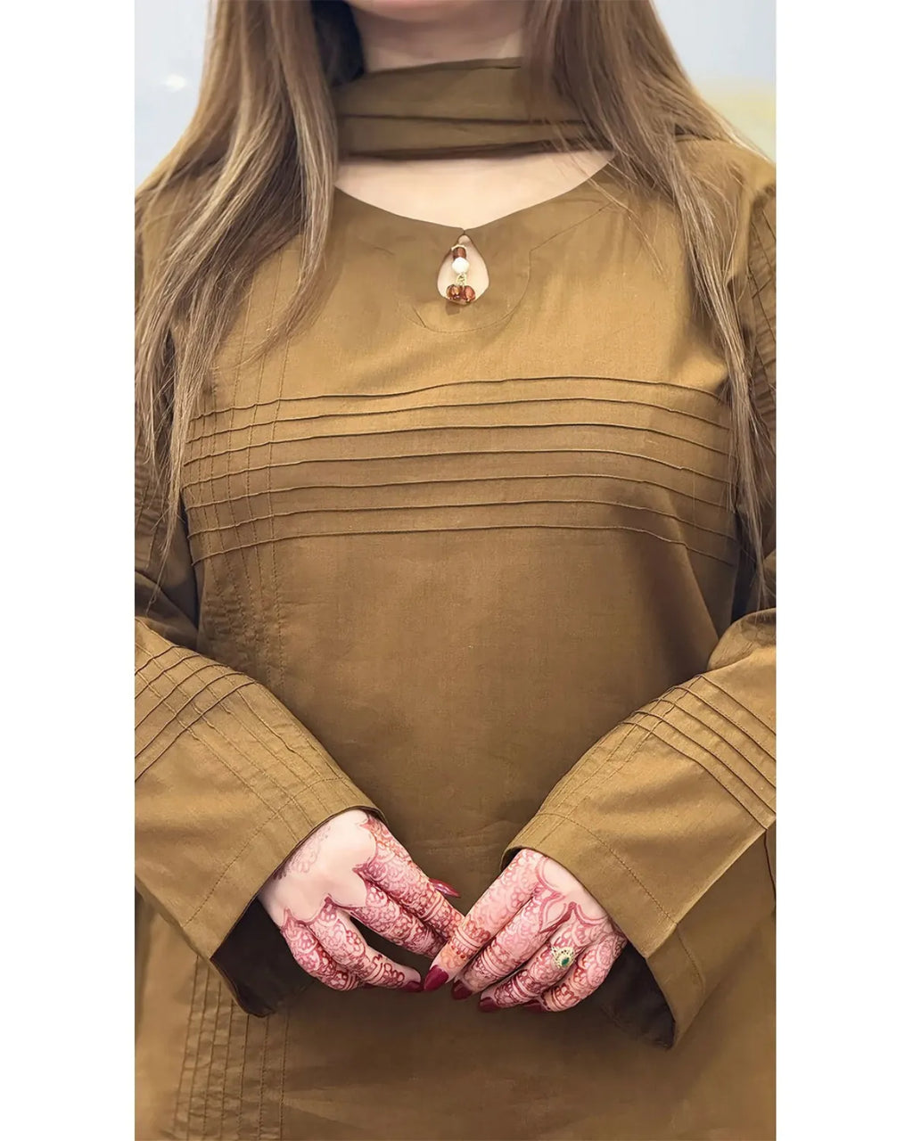 A woman wearing a brown long-sleeve tunic top with horizontal pleated details across the chest and cuffs, styled with a small pendant necklace. The photo is cropped from the neck down, showing her hands decorated with intricate red henna patterns.