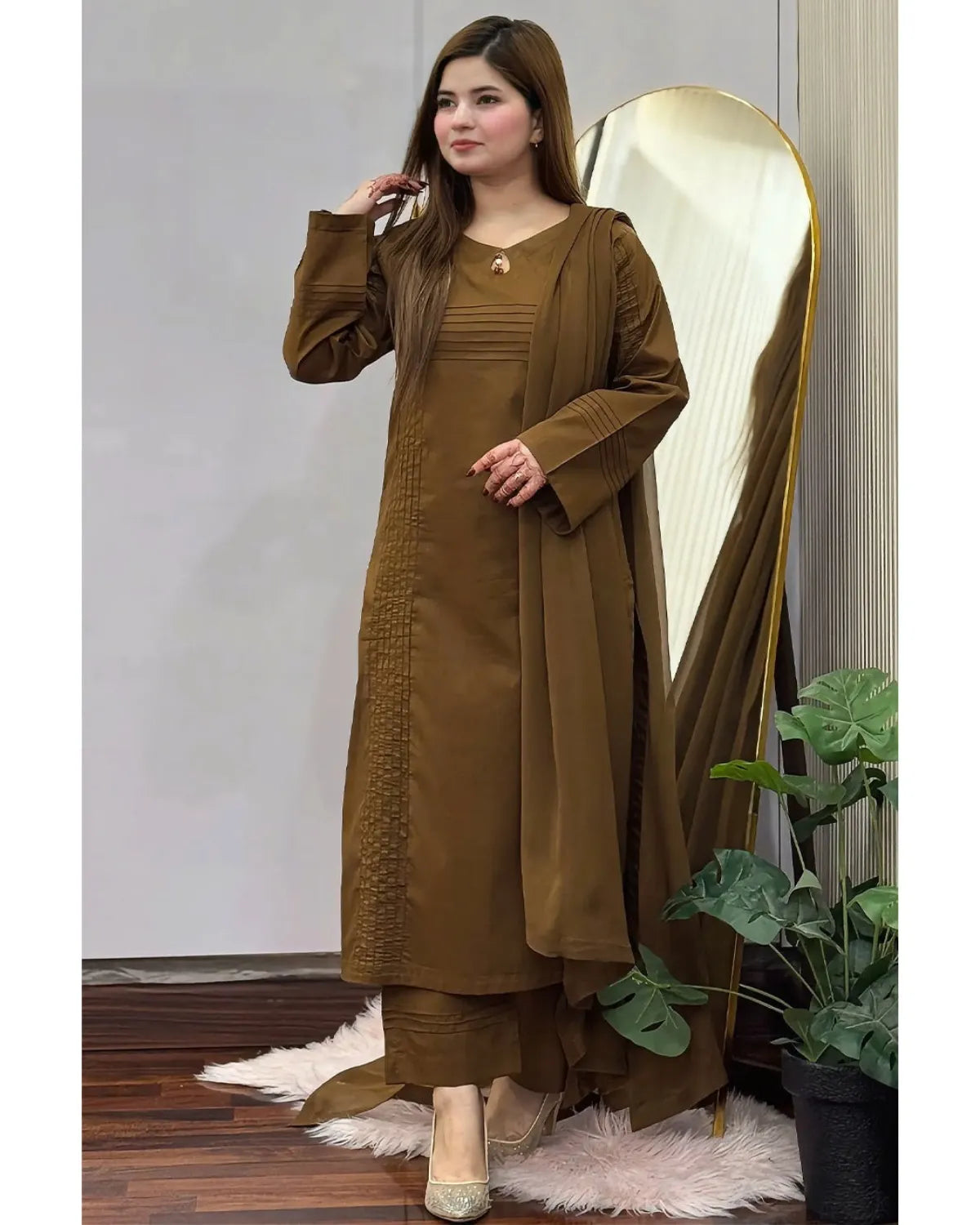 A woman models a brown traditional outfit consisting of a long-sleeved kurta with matching pants and a draped outer layer, standing in front of a large gold-framed mirror in an indoor setting.