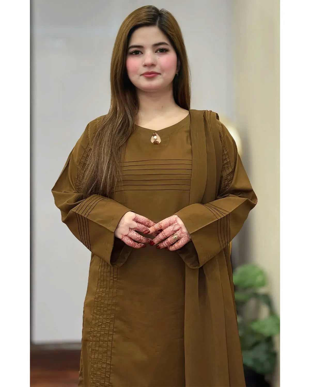 A woman wearing a brown long-sleeved dress with pleated details and a matching draped shawl, standing against a softly blurred indoor background.