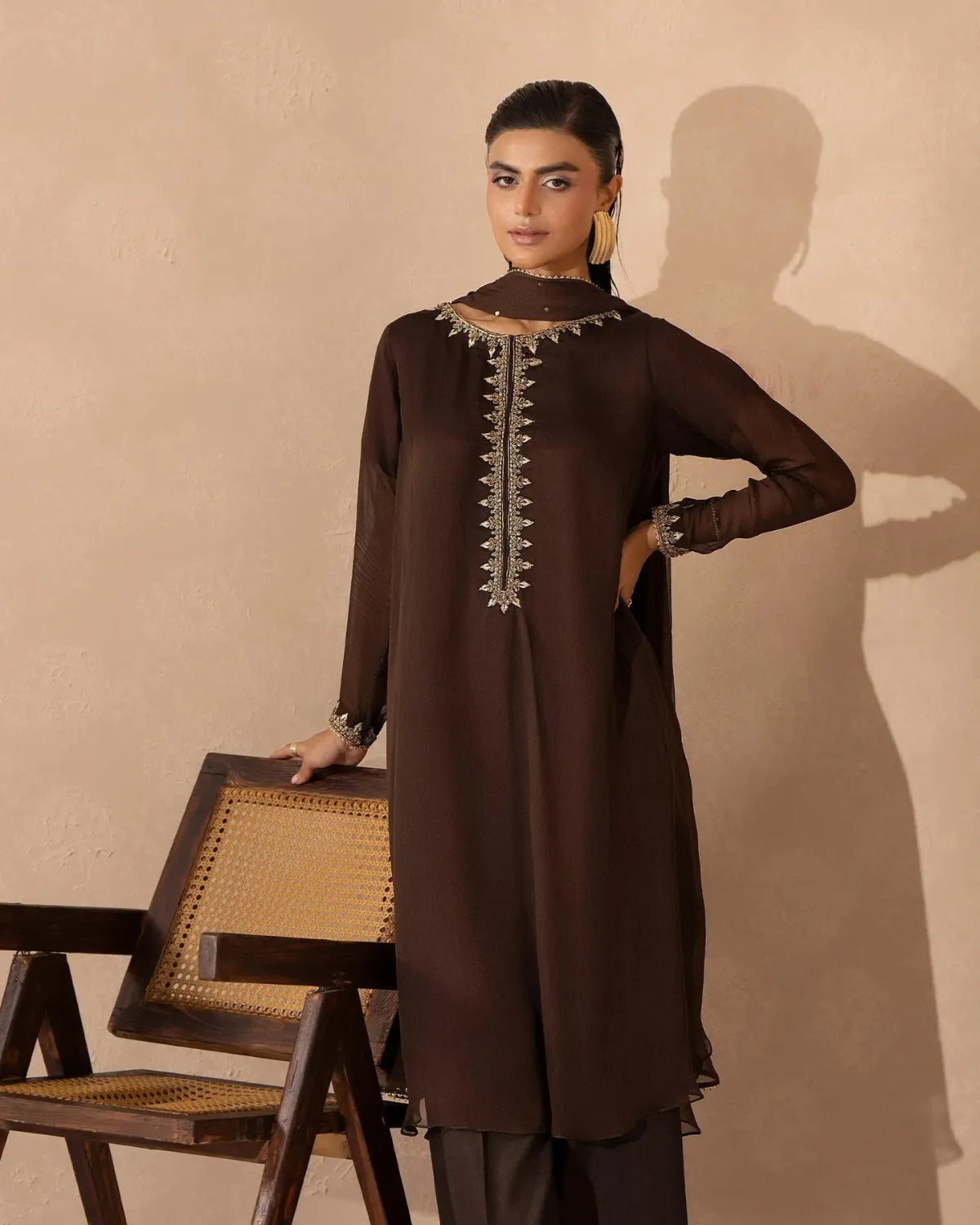A woman models a dark brown long kurta with silver embroidery on the neckline and front placket, paired with matching dark brown trousers, standing next to a wooden chair against a textured beige wall.