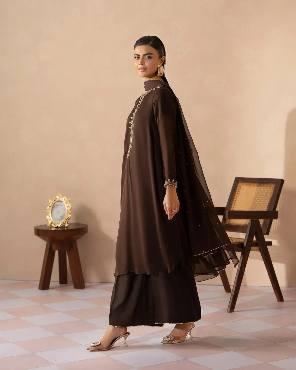 A woman models a dark brown traditional outfit consisting of a long-sleeved kurta with gold embroidery, matching wide-leg pants, and a sheer embroidered dupatta, set against a beige wall with a wooden chair and side table.