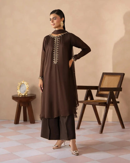 A woman models a dark brown traditional Indian salwar kameez suit with a matching dupatta, featuring gold embroidery on the neckline, in a styled studio setting with a wooden chair and side table.