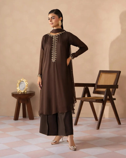 A woman models a dark brown traditional Indian salwar kameez suit with a matching dupatta, featuring gold embroidery on the neckline, in a styled studio setting with a wooden chair and side table.