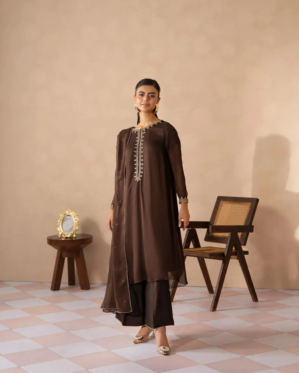 A woman models a dark brown traditional kurta set with matching pants in a styled studio setting. The kurta features gold embroidery along the neckline and sleeve cuffs, paired with a long sheer overlay.