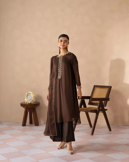 A woman models a dark brown traditional kurta set with matching pants in a styled studio setting. The kurta features gold embroidery along the neckline and sleeve cuffs, paired with a long sheer overlay.