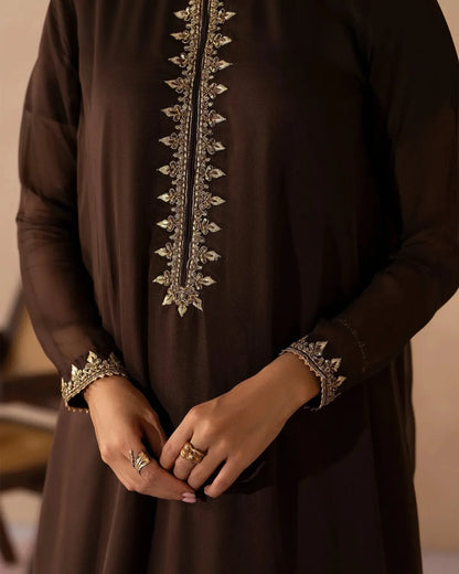 A close-up of a person wearing a dark brown long-sleeve tunic or dress with ornate metallic embroidery down the front placket and on the cuffs, with hands clasped in front.