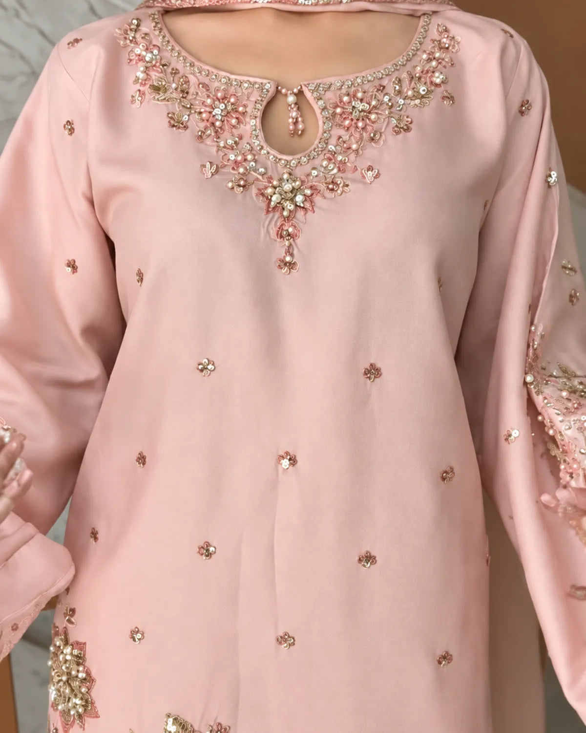 A light pink traditional long-sleeve tunic with ornate floral embroidery and beadwork around the neckline, decorated with scattered small metallic embellishments, shown on a model.