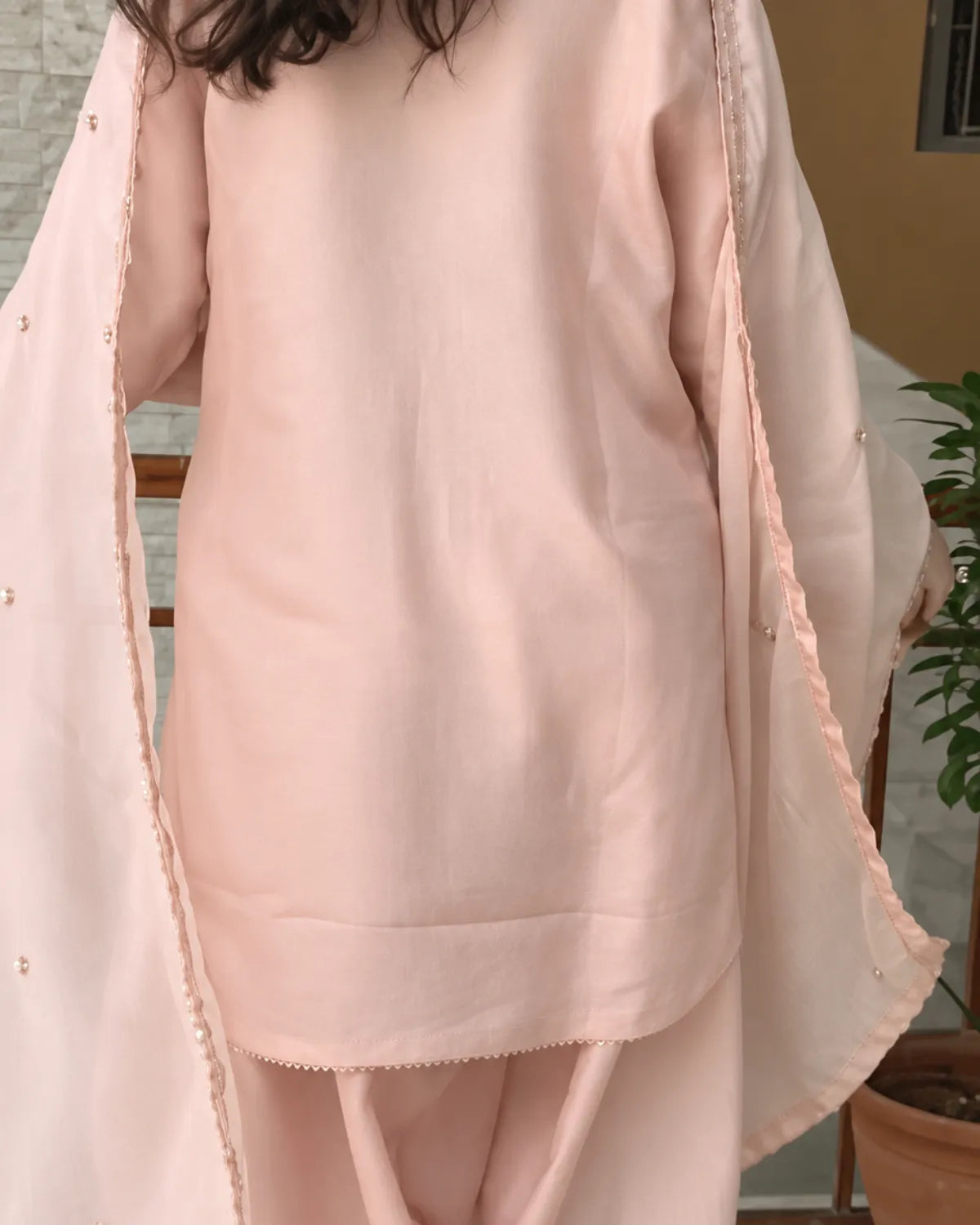 A person wearing a light blush pink traditional outfit with a matching draped scarf, photographed indoors with a neutral wall and potted plant in the background.
