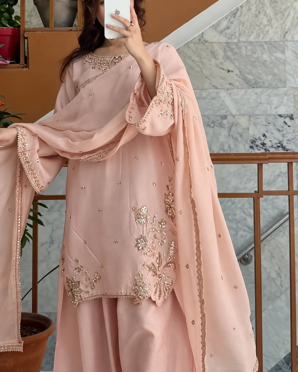 A woman takes a mirror selfie wearing a light pink traditional outfit with gold embroidery and a matching dupatta in an indoor setting with marble stairs and wooden railings.