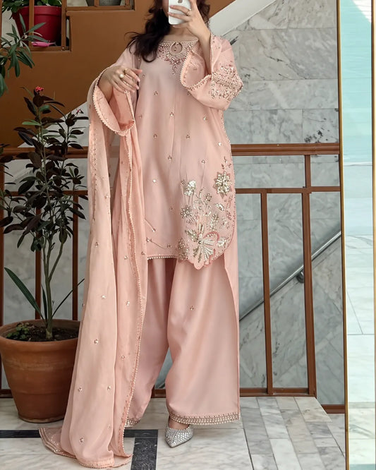 A woman modeling a light pink traditional three-piece outfit with wide-leg pants, a matching long dupatta, and intricate floral embroidery on the kurta, standing in front of a marble staircase.