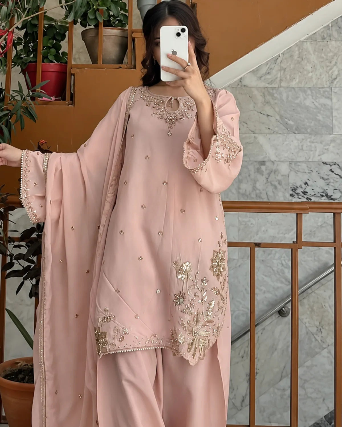 A woman takes a mirror selfie wearing a light pink traditional outfit with intricate gold and silver embroidery on the kurta and dupatta, set against an indoor staircase background.