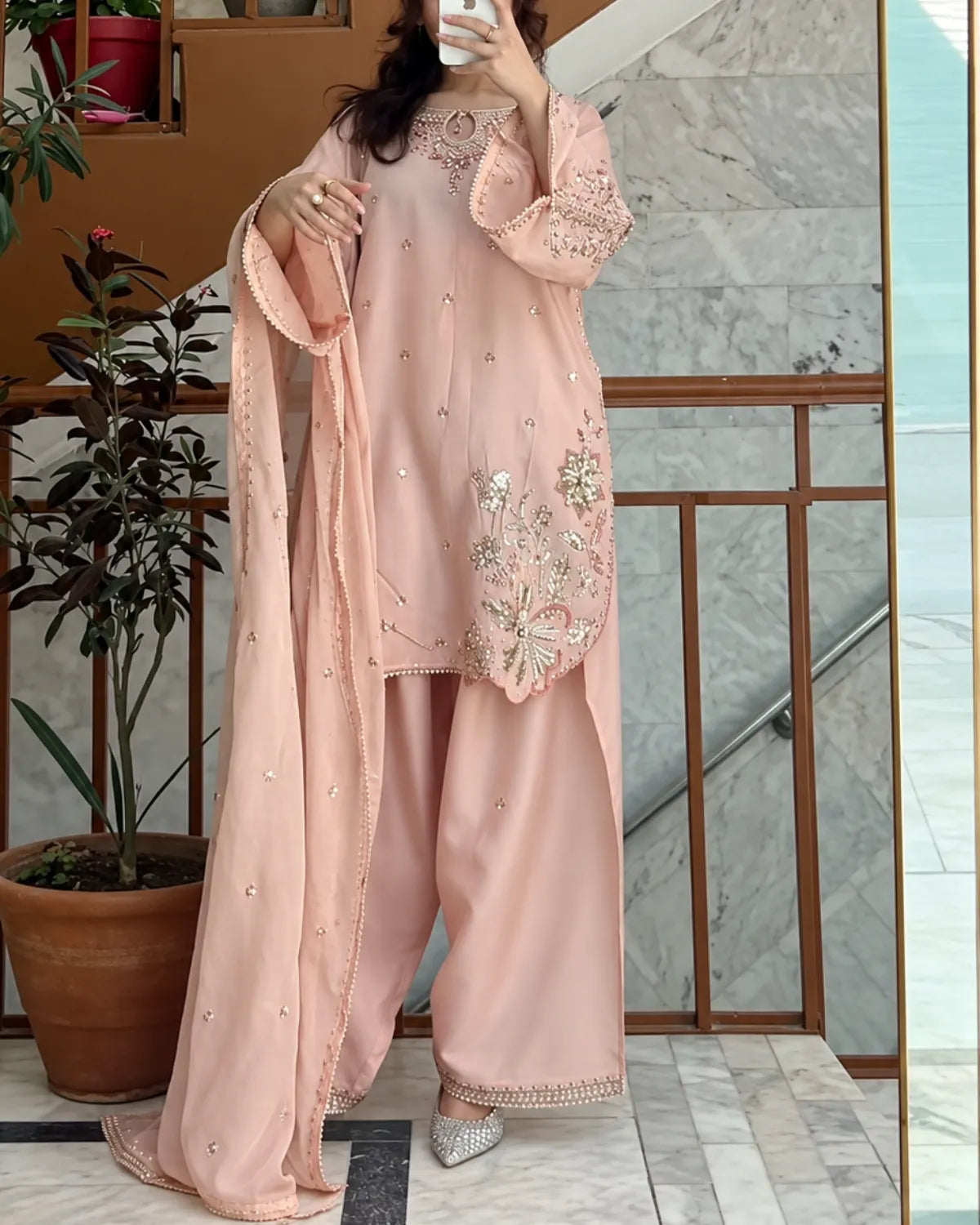 A woman takes a mirror selfie wearing a light peach-pink three-piece traditional outfit with matching pants and dupatta, featuring silver and gold embroidery on the kurta and border details, set against an indoor staircase background.