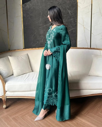 A woman models a long emerald green satin abaya with intricate silver embroidery and lace details, standing in a modern living room setting with a cream-colored sofa and marble walls.