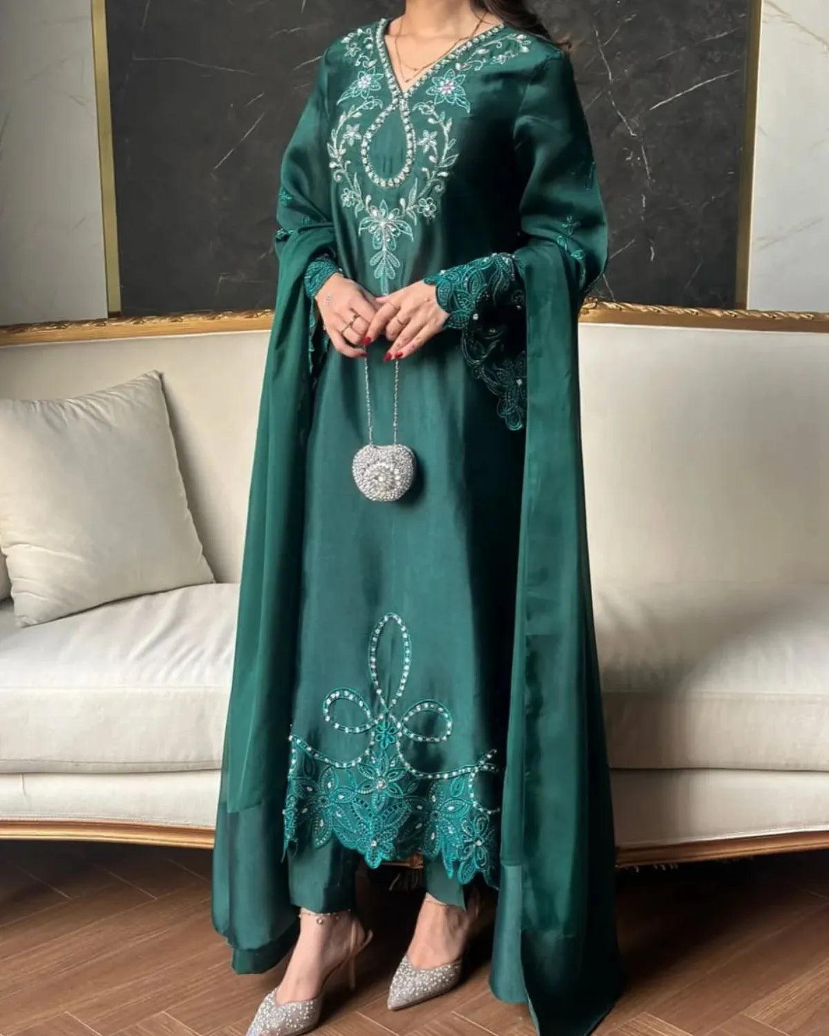 A woman models a dark teal green traditional long dress with intricate silver and white embroidery on the bodice and hem, paired with a matching long cape and holding a silver beaded purse.