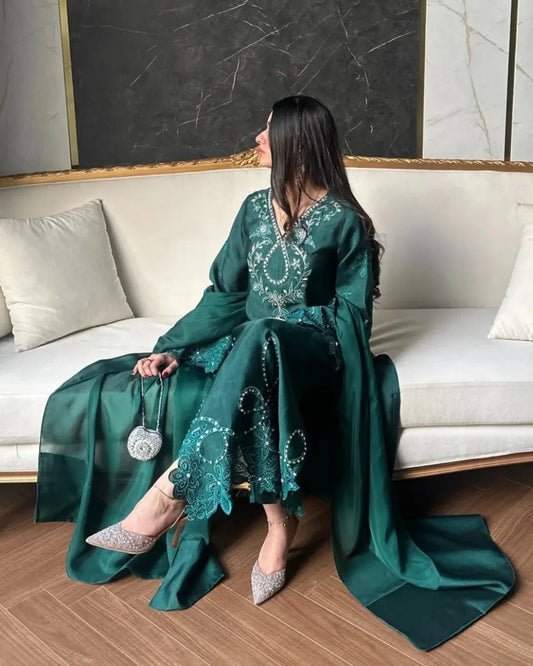 A woman sits on a cream-colored sofa wearing a flowing emerald green traditional outfit with intricate silver embroidery and long draped sleeves. She is holding a small silver handbag and wearing sparkly silver pointed-toe heels, set against a modern interior with a marble accent wall and wood flooring.