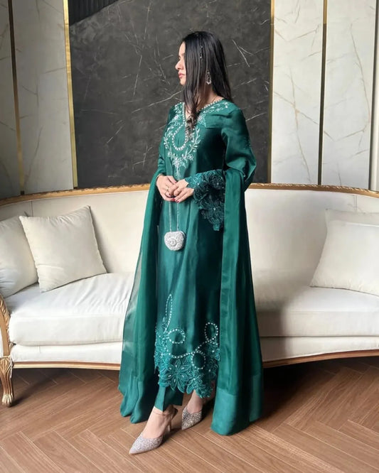 A woman models a long teal green traditional dress with silver embroidery and lace details, standing in front of a cream-colored sofa in a modern living room setting.
