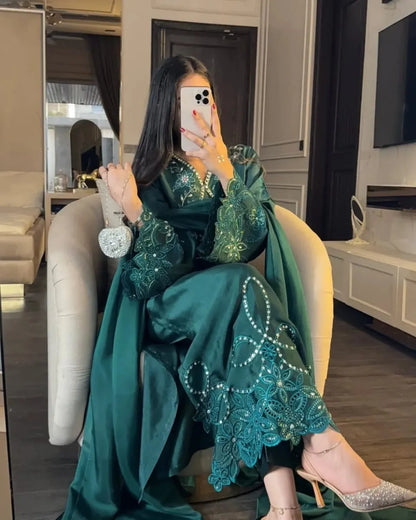 A woman takes a mirror selfie wearing a dark green embroidered traditional outfit with intricate beadwork and lace detailing, paired with sparkly silver heels in a modern living room setting.