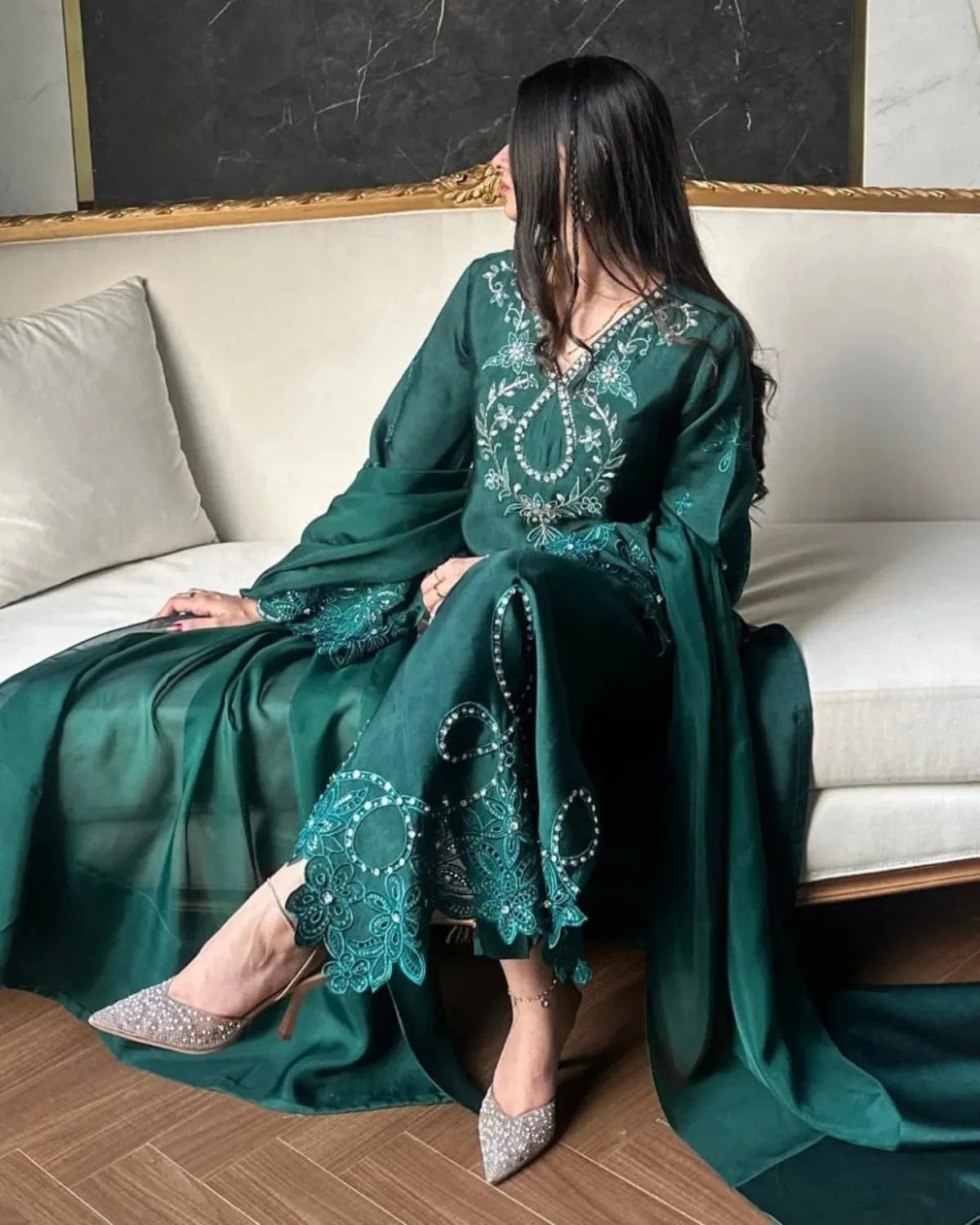 A woman sits on a cream-colored sofa wearing a flowing emerald green traditional dress with intricate silver embroidery and matching embroidered sleeves. She is wearing sparkly silver high heels, set against a background of a dark marble wall and wooden floor.