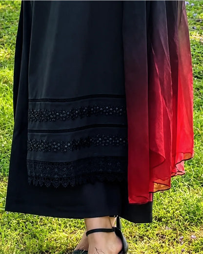 A close-up of a black dress or tunic with decorative embroidered and lace borders, paired with a sheer red and black gradient drape, shown outdoors on a green grass background.