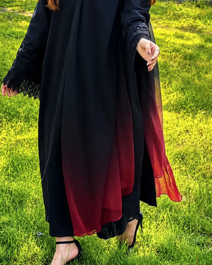 A person outdoors on grass wearing a long black abaya or dress with a deep red ombré gradient near the hem and sleeves, plus black lace trim at the cuffs.