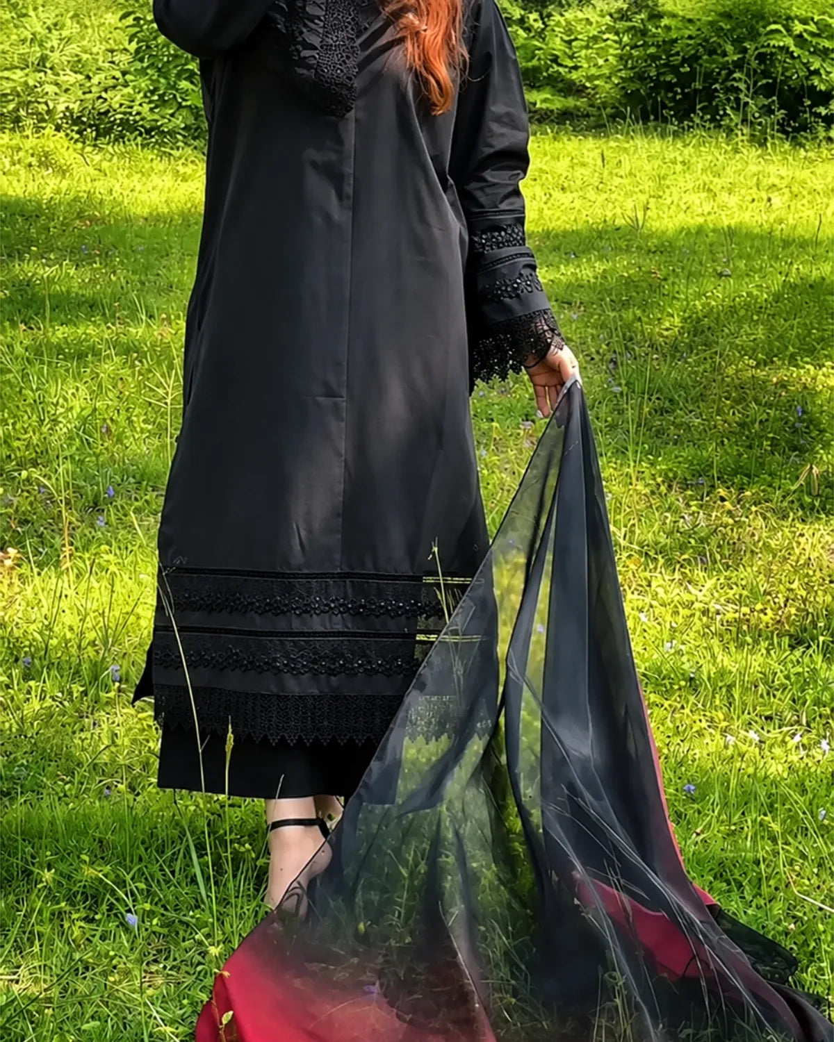A person standing in a grassy outdoor setting wearing a long black dress or tunic with lace trim and holding a sheer black dupatta or scarf with a red border.