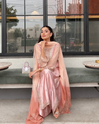 A woman wearing a light pink traditional outfit with a matching sheer dupatta sits on a bench in front of a modern building with large black-framed windows.